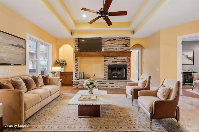 living room with ceiling fan, a stone fireplace, and a raised ceiling