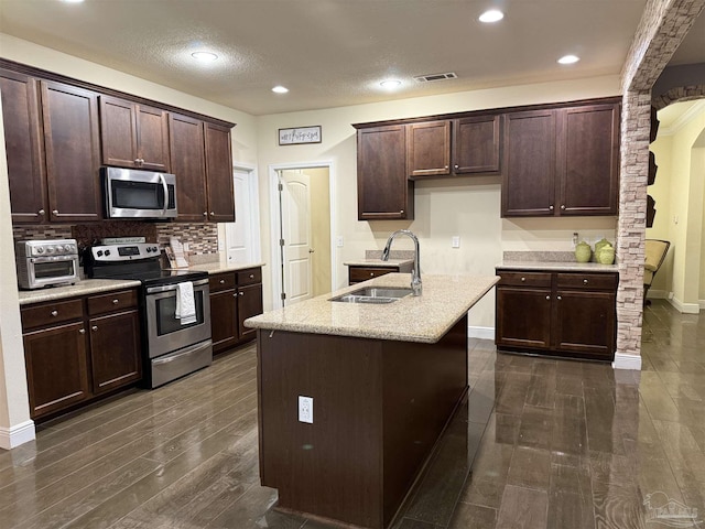 kitchen with dark wood-style flooring, stainless steel appliances, visible vents, a sink, and an island with sink