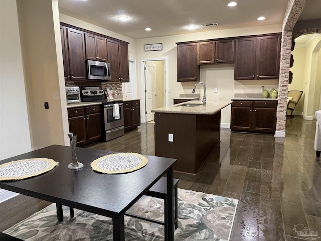 kitchen with arched walkways, dark wood finished floors, an island with sink, appliances with stainless steel finishes, and a sink