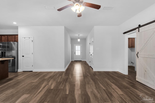 unfurnished living room with a barn door, a ceiling fan, dark wood finished floors, baseboards, and visible vents