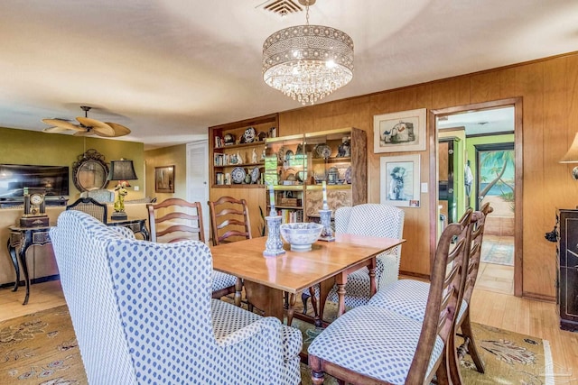 dining room with ceiling fan with notable chandelier, light hardwood / wood-style floors, and wooden walls