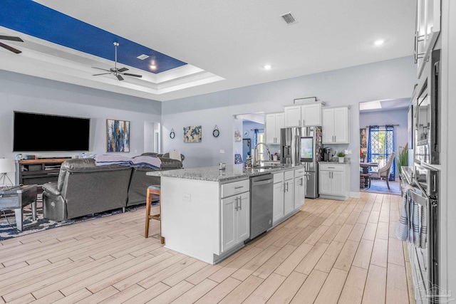 kitchen with white cabinets, ceiling fan, a tray ceiling, stainless steel appliances, and a center island with sink