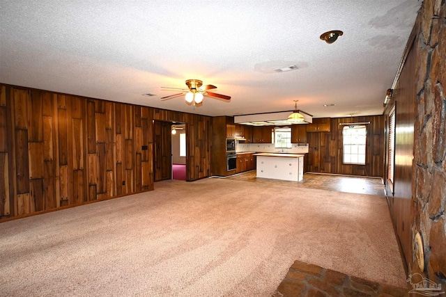 unfurnished living room with ceiling fan, light colored carpet, wooden walls, and a textured ceiling