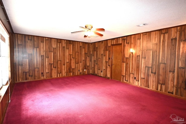 unfurnished room with a textured ceiling, wood walls, ceiling fan, and dark carpet