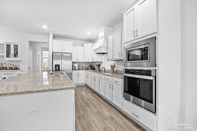 kitchen featuring stainless steel appliances, a sink, white cabinetry, custom range hood, and light stone countertops