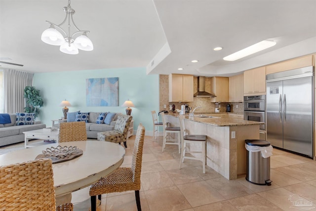 kitchen featuring wall chimney exhaust hood, stainless steel appliances, a notable chandelier, decorative light fixtures, and light brown cabinetry