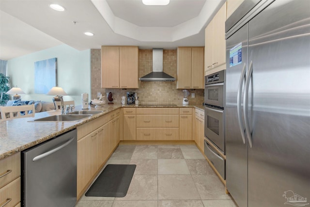 kitchen with light brown cabinets, wall chimney exhaust hood, a raised ceiling, decorative backsplash, and appliances with stainless steel finishes