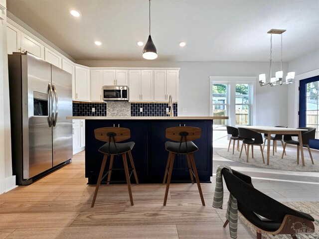 kitchen with appliances with stainless steel finishes, white cabinets, decorative light fixtures, light wood-type flooring, and an island with sink