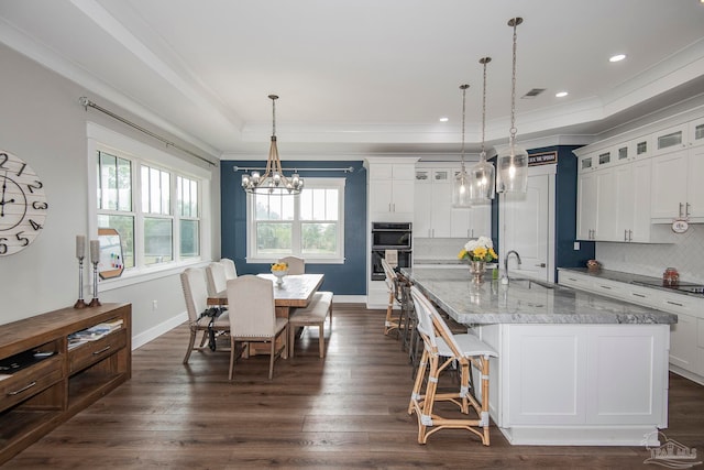 kitchen with dark wood-type flooring, hanging light fixtures, backsplash, double oven, and a kitchen island with sink