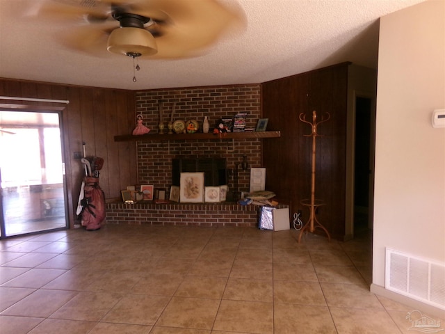 tiled living room with a brick fireplace, a textured ceiling, ceiling fan, and wood walls