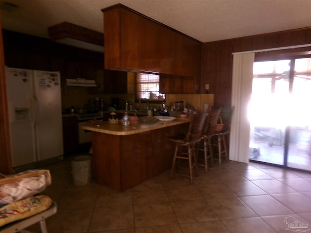 kitchen with wooden walls, white refrigerator with ice dispenser, range, light tile patterned floors, and kitchen peninsula