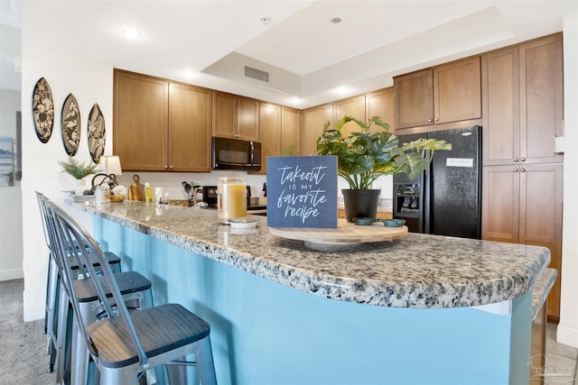 kitchen featuring a breakfast bar, a tray ceiling, light stone countertops, black appliances, and kitchen peninsula