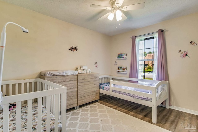 bedroom with ceiling fan, a textured ceiling, a crib, and dark wood-type flooring