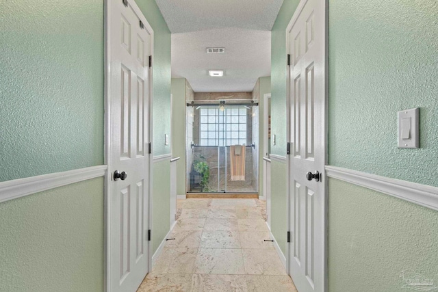 hallway with a textured wall, visible vents, baseboards, and stone tile flooring