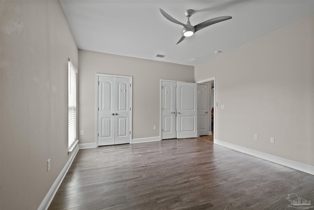 unfurnished bedroom featuring ceiling fan, dark wood-type flooring, visible vents, baseboards, and multiple closets