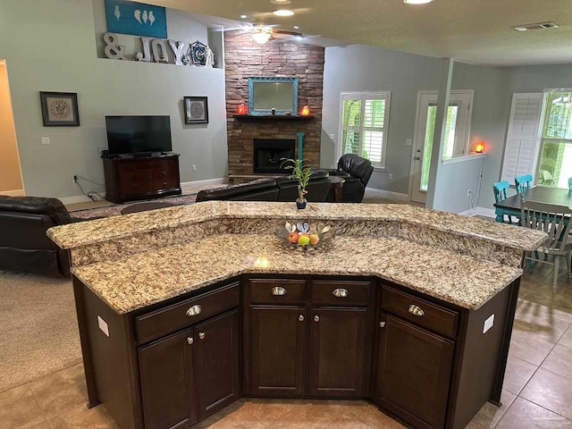 kitchen with open floor plan, a ceiling fan, light stone counters, dark brown cabinets, and a stone fireplace