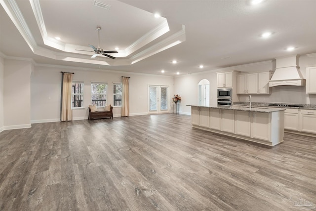 kitchen featuring premium range hood, stainless steel appliances, light stone counters, a tray ceiling, and a center island with sink