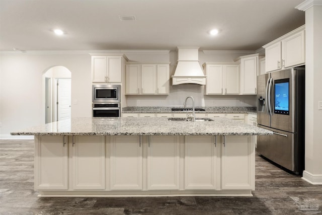 kitchen featuring appliances with stainless steel finishes, light stone countertops, white cabinets, a center island with sink, and custom exhaust hood