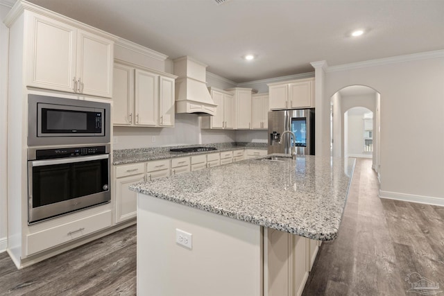 kitchen with custom exhaust hood, light stone counters, crown molding, appliances with stainless steel finishes, and an island with sink