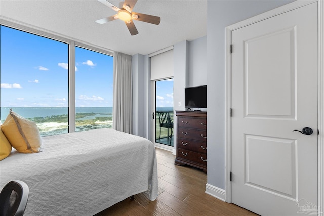 bedroom with a textured ceiling, expansive windows, dark wood-type flooring, a water view, and ceiling fan