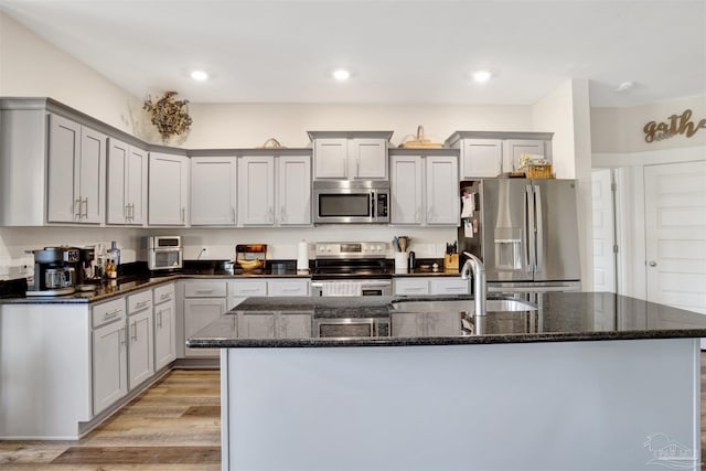 kitchen with dark stone countertops, light hardwood / wood-style floors, sink, a kitchen island with sink, and appliances with stainless steel finishes