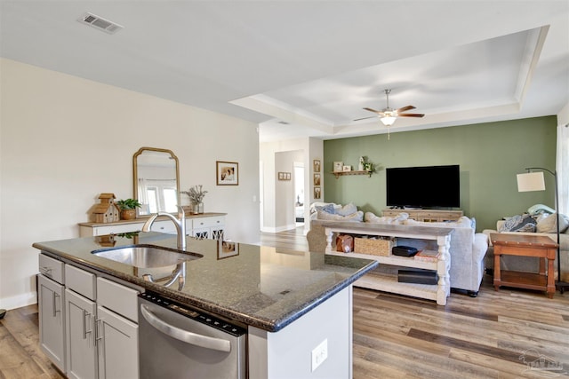 kitchen with dark stone countertops, stainless steel dishwasher, a raised ceiling, and an island with sink