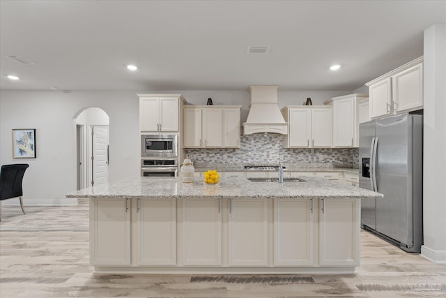 kitchen with light stone counters, custom exhaust hood, stainless steel appliances, and a center island with sink