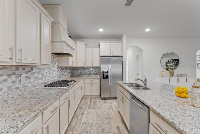 kitchen with premium range hood, appliances with stainless steel finishes, sink, light stone counters, and light wood-type flooring