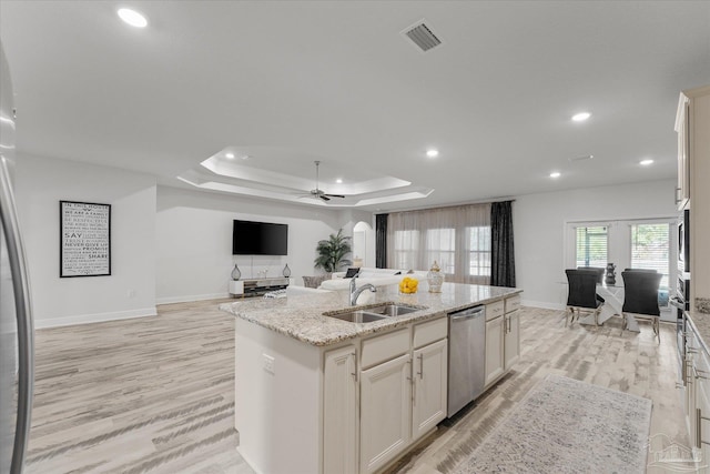 kitchen with sink, a kitchen island with sink, stainless steel dishwasher, light stone counters, and a raised ceiling