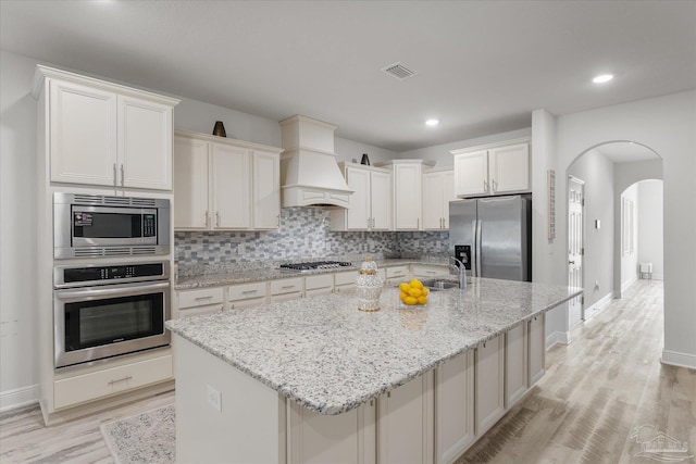 kitchen with light stone counters, stainless steel appliances, a kitchen island with sink, and custom exhaust hood