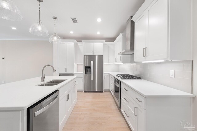 kitchen with light wood-type flooring, appliances with stainless steel finishes, wall chimney exhaust hood, decorative backsplash, and decorative light fixtures