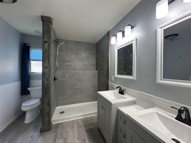 bathroom featuring a wainscoted wall, toilet, wood finished floors, tiled shower, and a textured ceiling