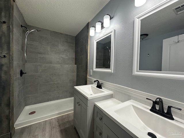 full bathroom with a textured ceiling, tiled shower, wood finished floors, two vanities, and a wainscoted wall