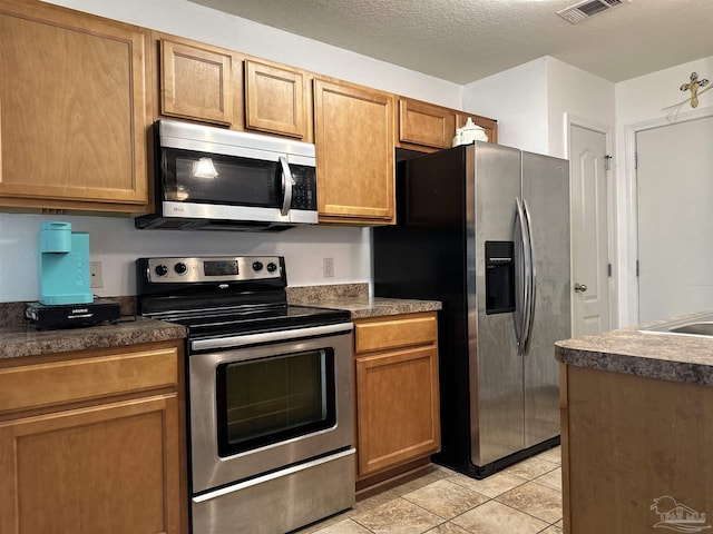 kitchen with appliances with stainless steel finishes, light tile patterned floors, dark countertops, and brown cabinetry