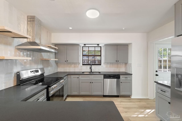 kitchen featuring dark countertops, wall chimney range hood, gray cabinets, stainless steel appliances, and a sink