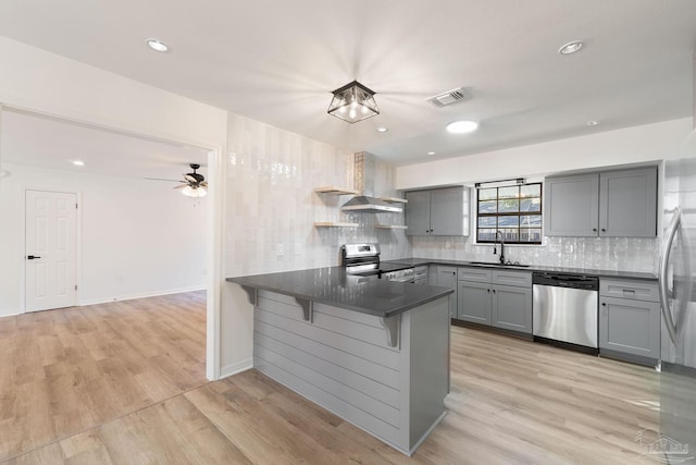 kitchen with gray cabinetry, a sink, stainless steel appliances, a peninsula, and wall chimney range hood