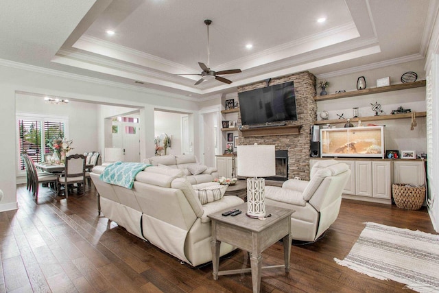 living area with a tray ceiling, ceiling fan, a fireplace, dark wood-style flooring, and crown molding