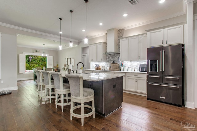 kitchen featuring high end fridge, wall chimney range hood, tasteful backsplash, a breakfast bar, and a kitchen island with sink