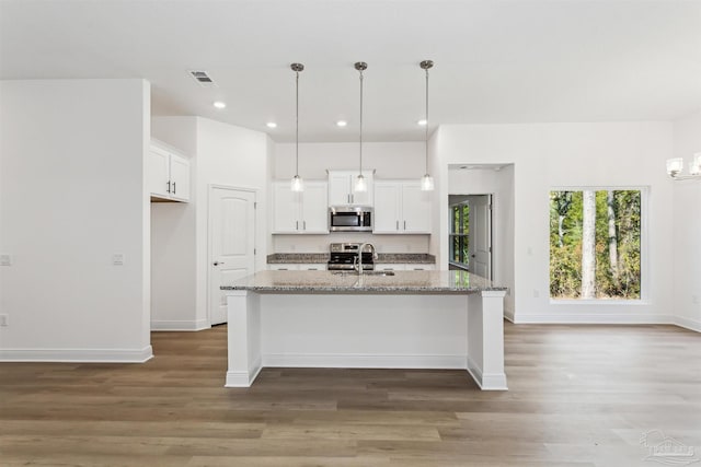 kitchen with light stone counters, white cabinetry, stainless steel appliances, hanging light fixtures, and a kitchen island with sink