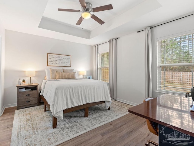 bedroom with ceiling fan, multiple windows, a raised ceiling, and light wood-type flooring