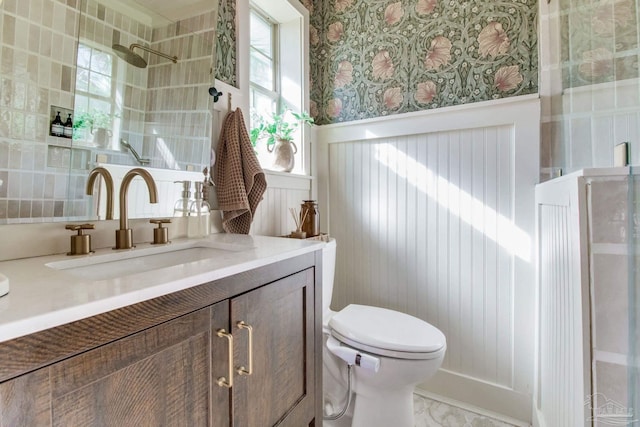 bathroom with wallpapered walls, a wainscoted wall, toilet, vanity, and a shower