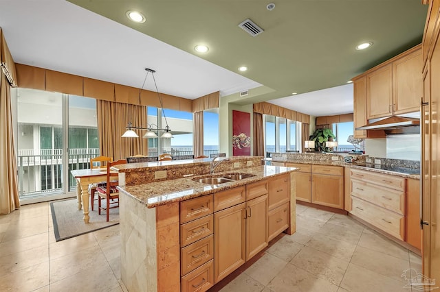 kitchen with sink, hanging light fixtures, light stone counters, an island with sink, and light brown cabinets