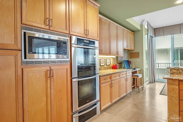 kitchen with light tile patterned flooring, light brown cabinetry, ornamental molding, stainless steel appliances, and light stone countertops