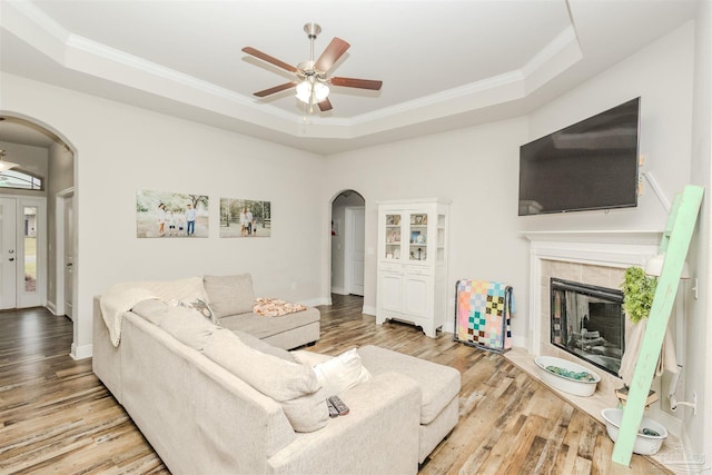 living area featuring arched walkways, light wood finished floors, ornamental molding, a tray ceiling, and a tiled fireplace