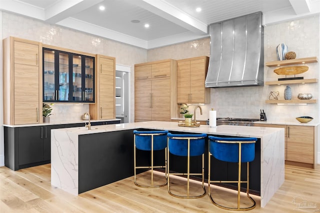 kitchen featuring beamed ceiling, a large island with sink, light wood-type flooring, wall chimney range hood, and a kitchen bar