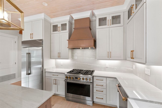 kitchen with high end appliances, wall chimney exhaust hood, tasteful backsplash, and white cabinets