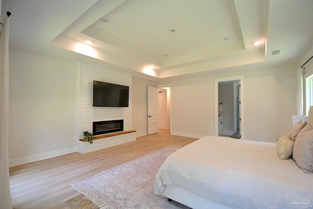 bedroom featuring a raised ceiling, a large fireplace, ensuite bathroom, and light wood-type flooring