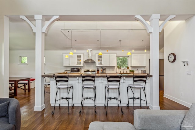 kitchen with appliances with stainless steel finishes, a kitchen breakfast bar, dark wood-type flooring, white cabinets, and wall chimney exhaust hood