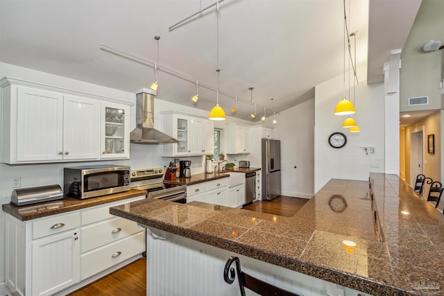 kitchen with appliances with stainless steel finishes, white cabinetry, wall chimney range hood, and pendant lighting