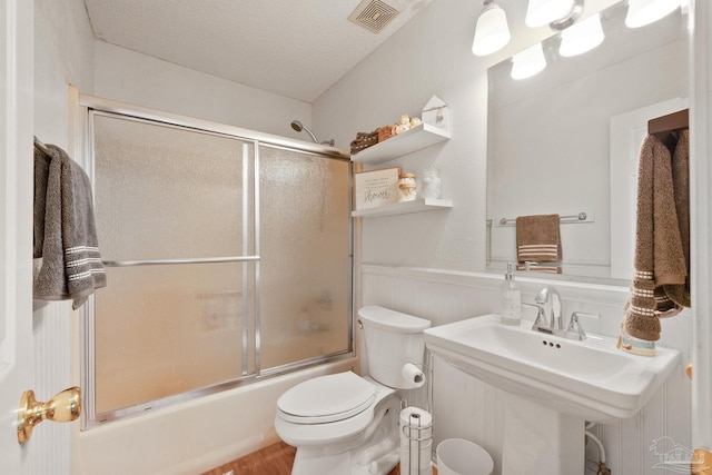 full bath featuring enclosed tub / shower combo, a textured ceiling, toilet, a wainscoted wall, and visible vents